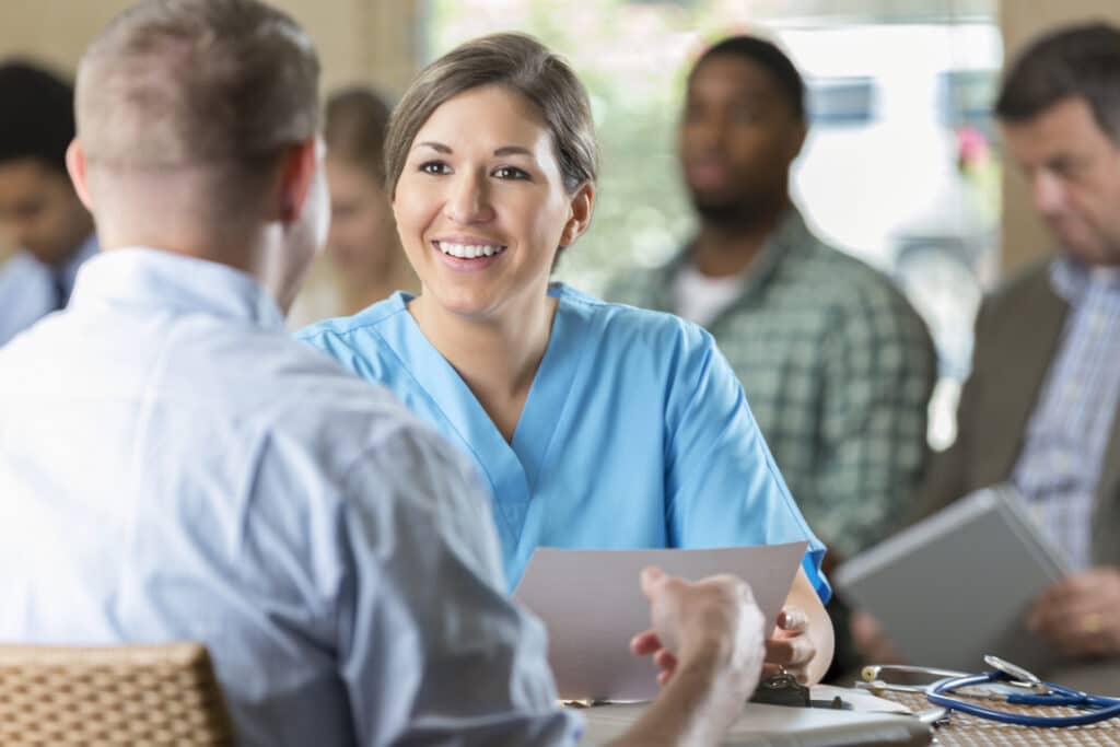 Mid adult woman is manager of at hospital. She is reading a resume and interviewing a potential nursing staff employee. Other candidates are waiting to be interviewed during hospital's staff recruitment event.