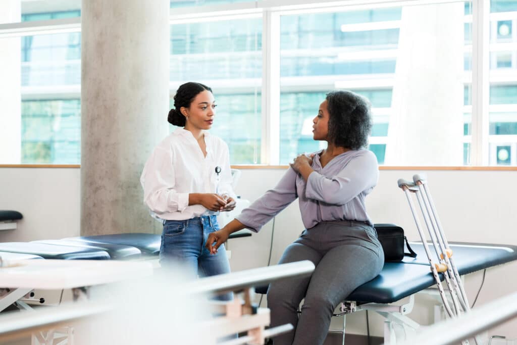 A mid adult woman touches her shoulder as she discusses pain or an injury with a female physical therapist. The patient is sitting on an exam table with crutches leaning against the table.